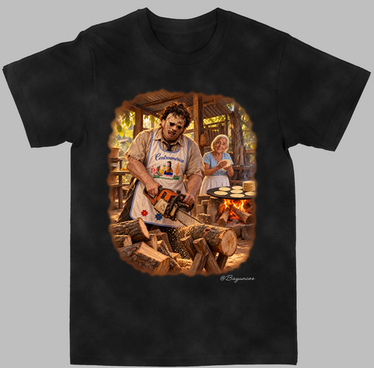 A black short-sleeve crewneck t-shirt featuring a large graphic print of Leatherface from the 'Psycho' movie, shown on a solid gray background cutting wood while a woman makes pupusas in the background